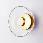 Close view of the clear glass dome and brushed brass centre disc of this Clérachic wall light on a pale wall.
