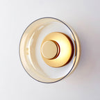 An amber glass version of this Clérachic wall light, shown unlit with a brass centre disc against a pale grey wall.