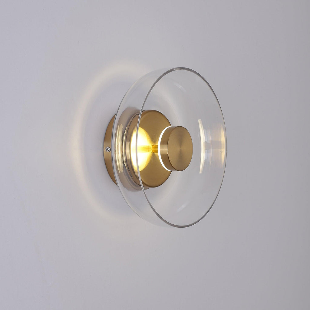 Angled view of a lit Clérachic wall light with a clear glass dome and brass fitting, its glow spilling out on a pale grey wall.