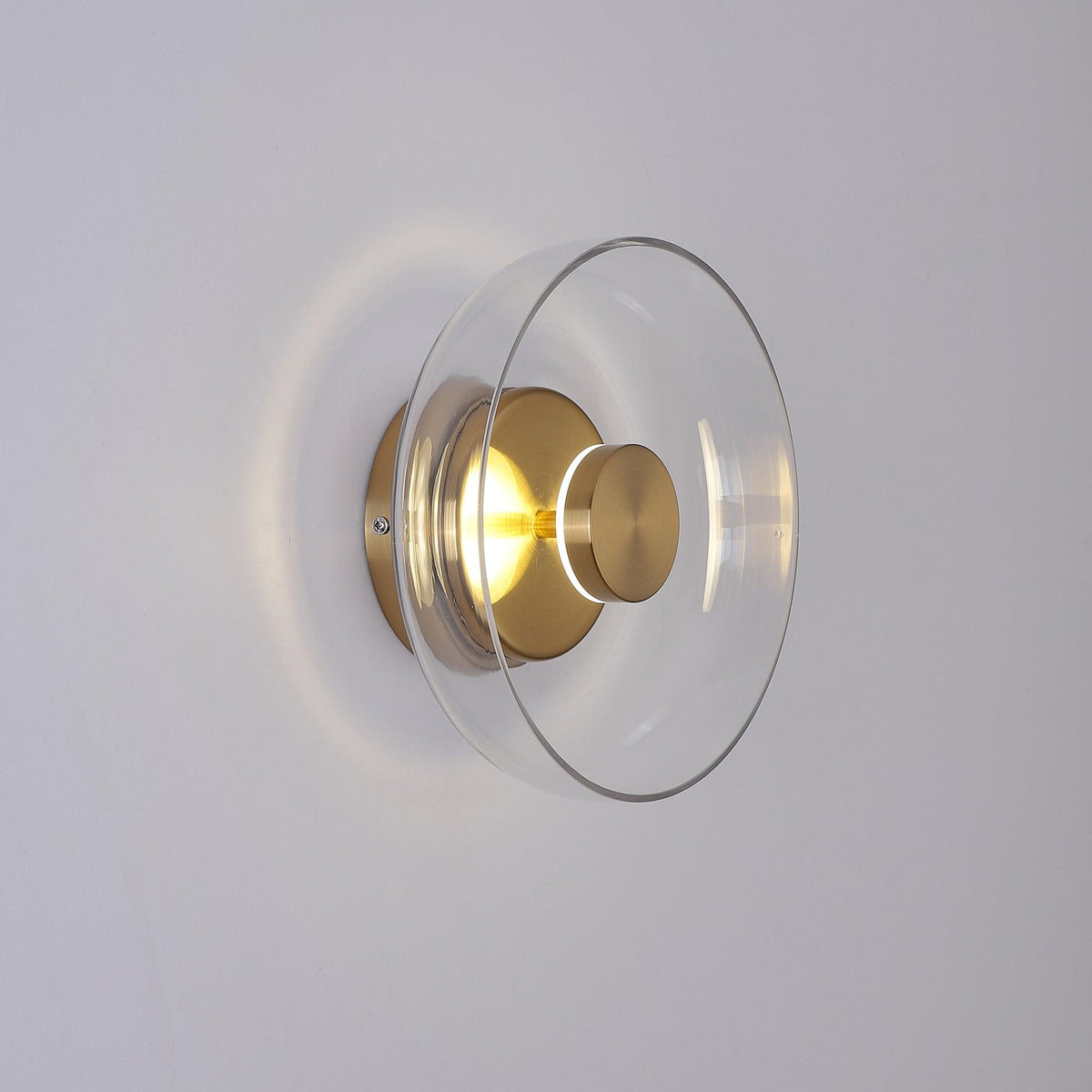 Angled view of a lit Clérachic wall light with a clear glass dome and brass fitting, its glow spilling out on a pale grey wall.