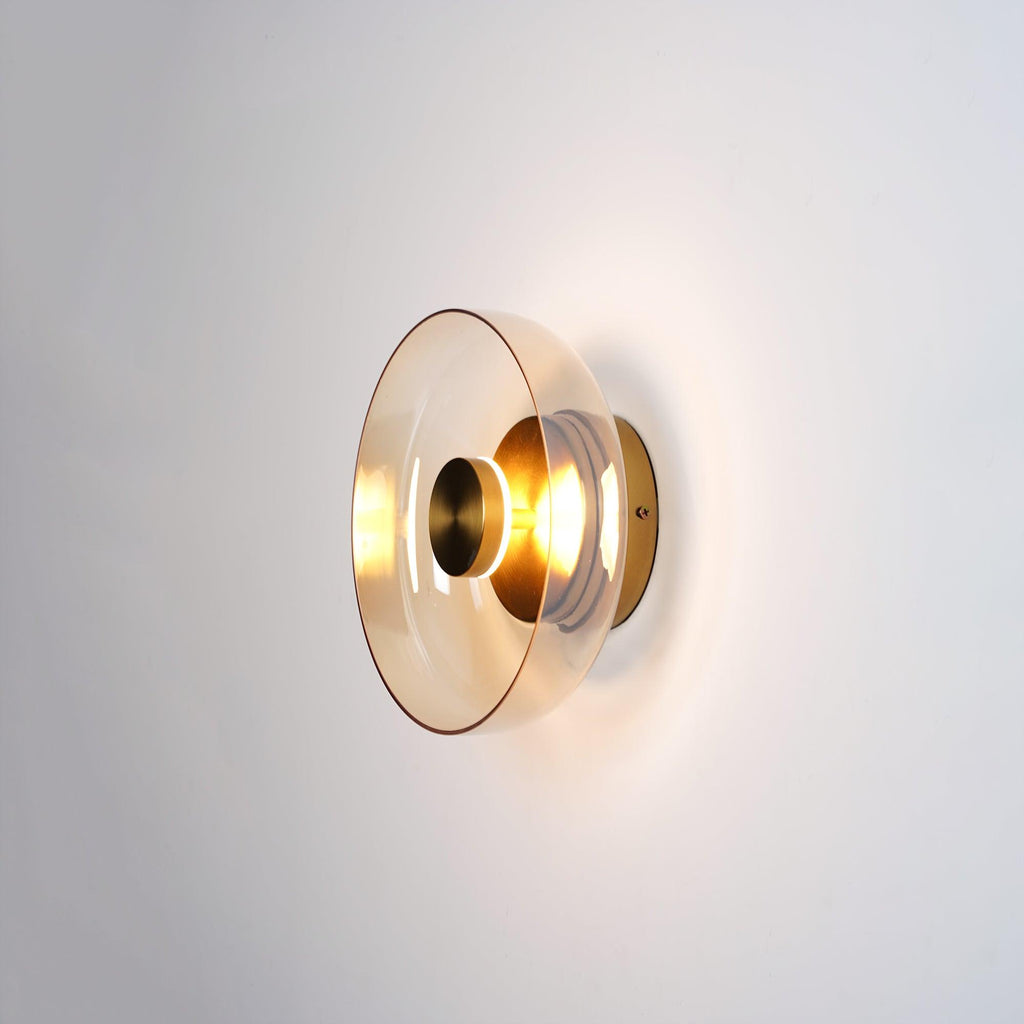 A Clérachic wall light with a smoky amber glass dome and a brass centre knob, glowing on a light wall.
