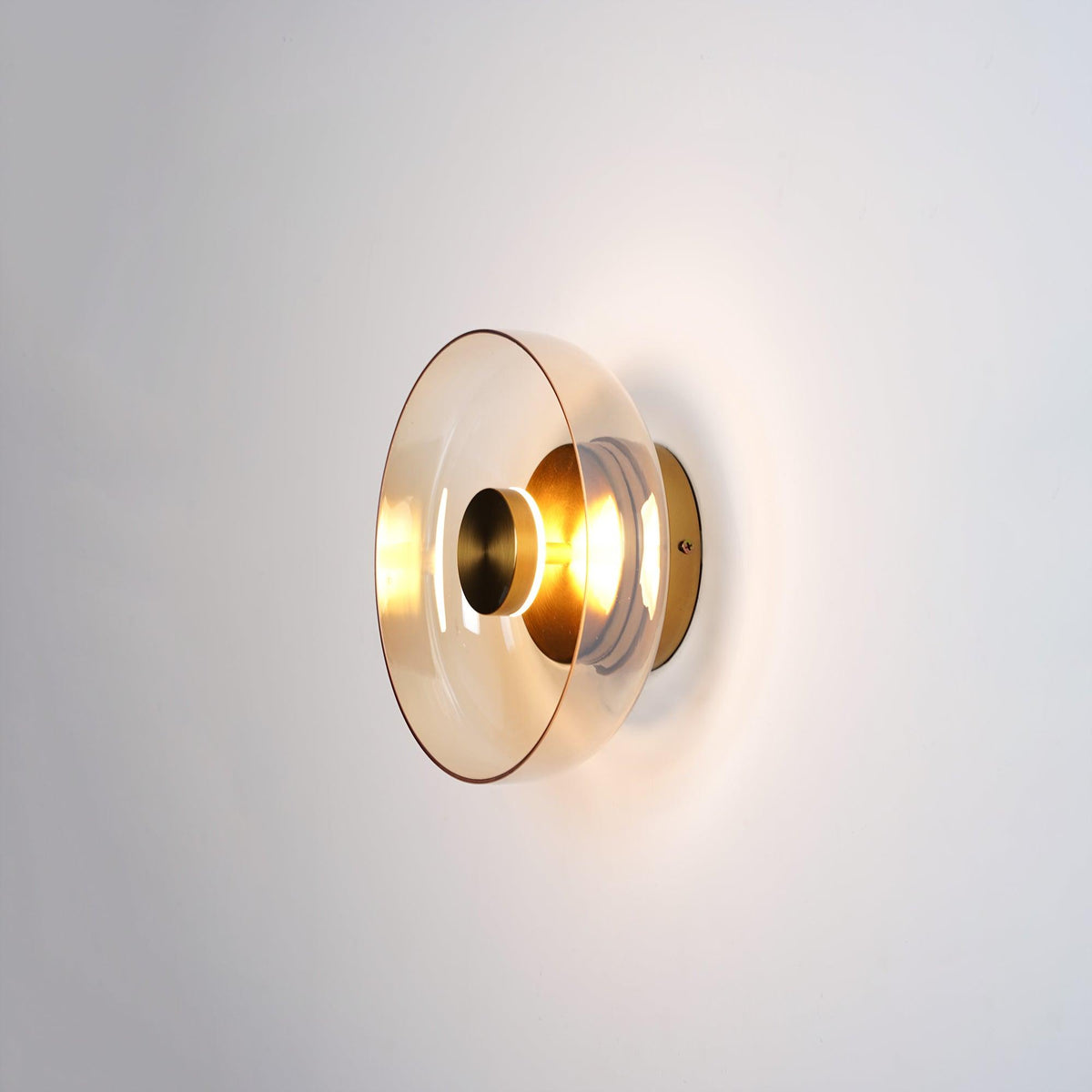 A Clérachic wall light with a smoky amber glass dome and a brass centre knob, glowing on a light wall.