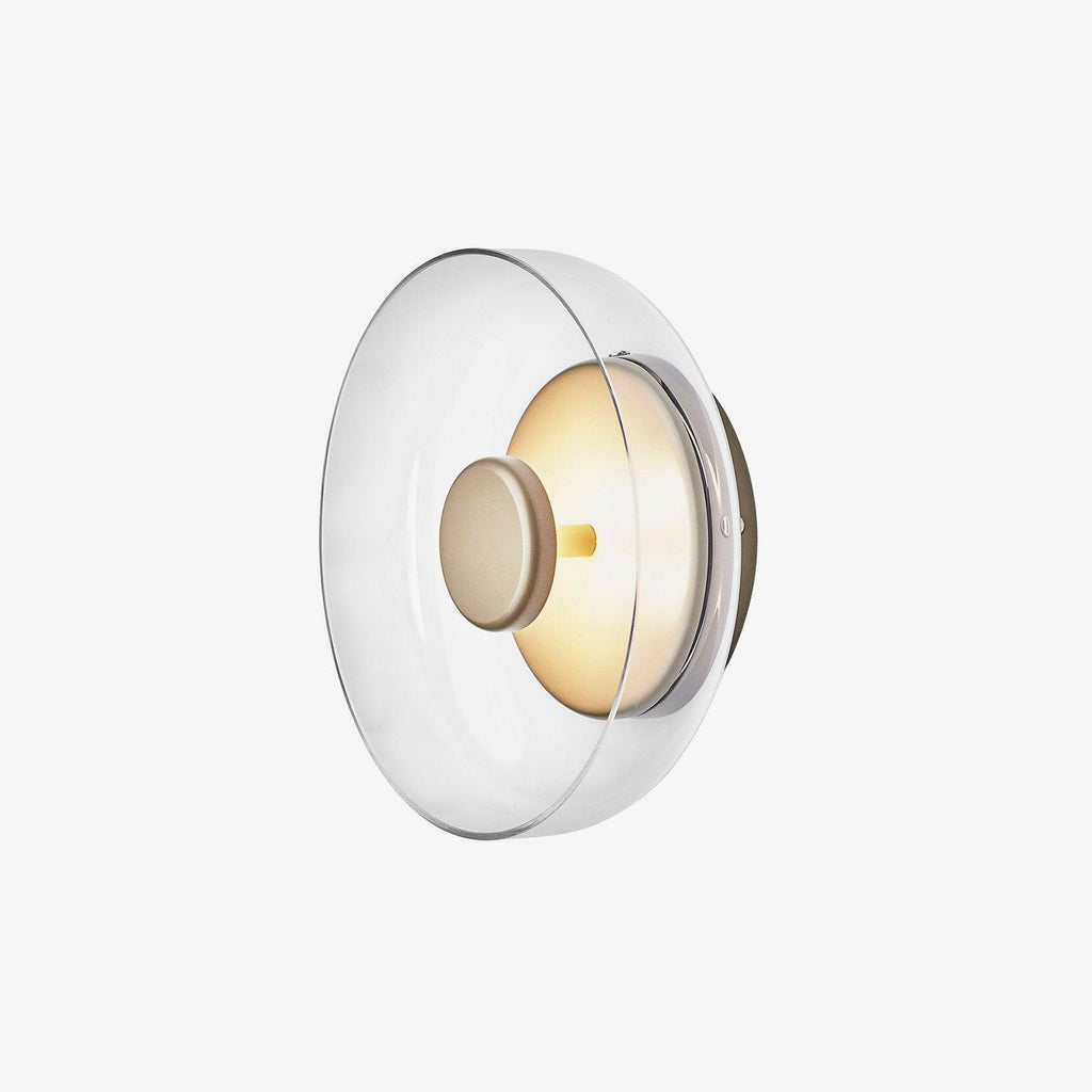 This Clérachic wall light on white, seen at an angle, has a wide clear glass dome and a brass wall plate with a soft glow.