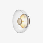 This Clérachic wall light on white, seen at an angle, has a wide clear glass dome and a brass wall plate with a soft glow.