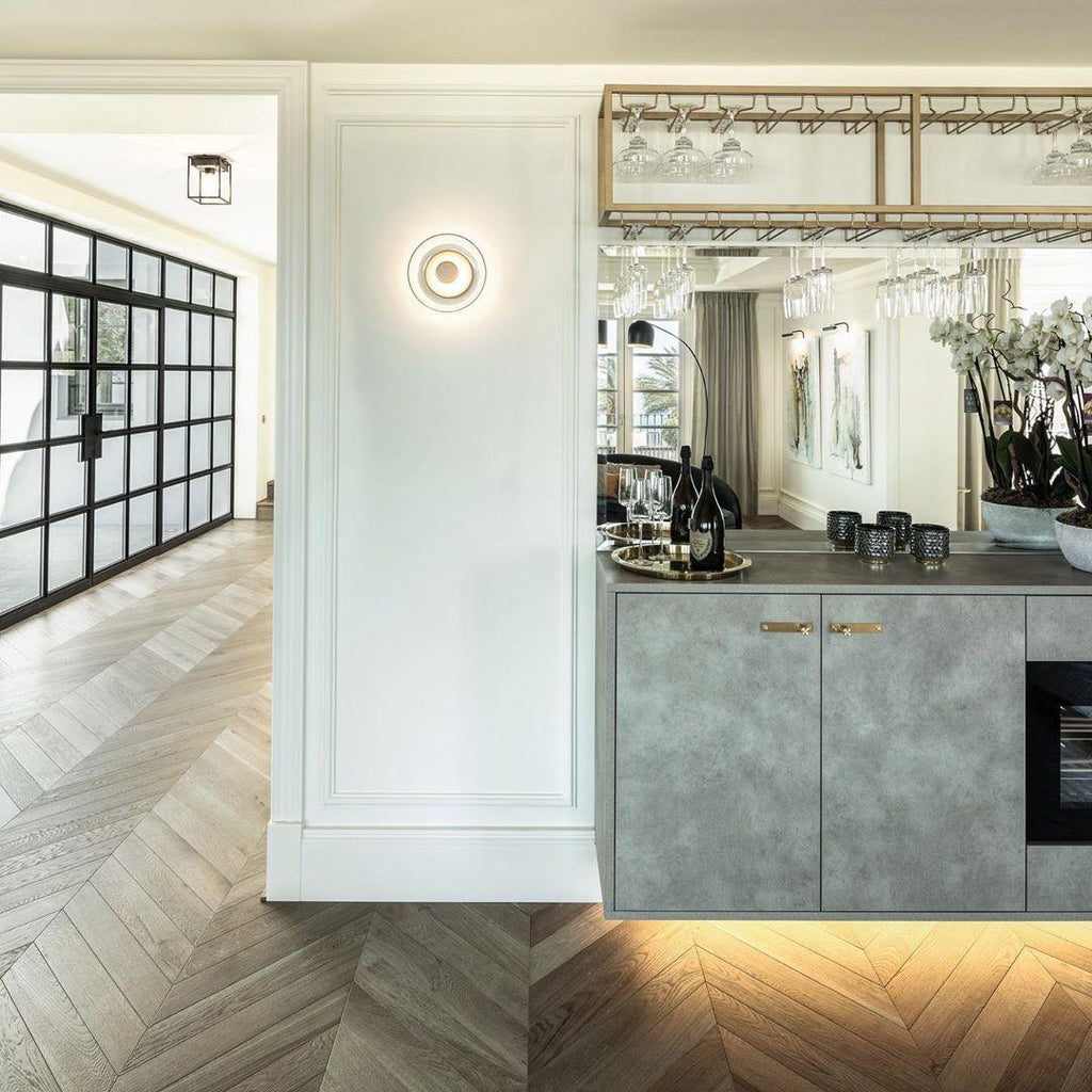 In an open kitchen with herringbone flooring and a glass-shelved bar, a Clérachic wall light glows softly on a white panelled wall.