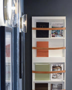 A Clérachic wall light with a clear glass dome seen edge-on beside framed pictures, above a shelf holding magazines on leather straps.