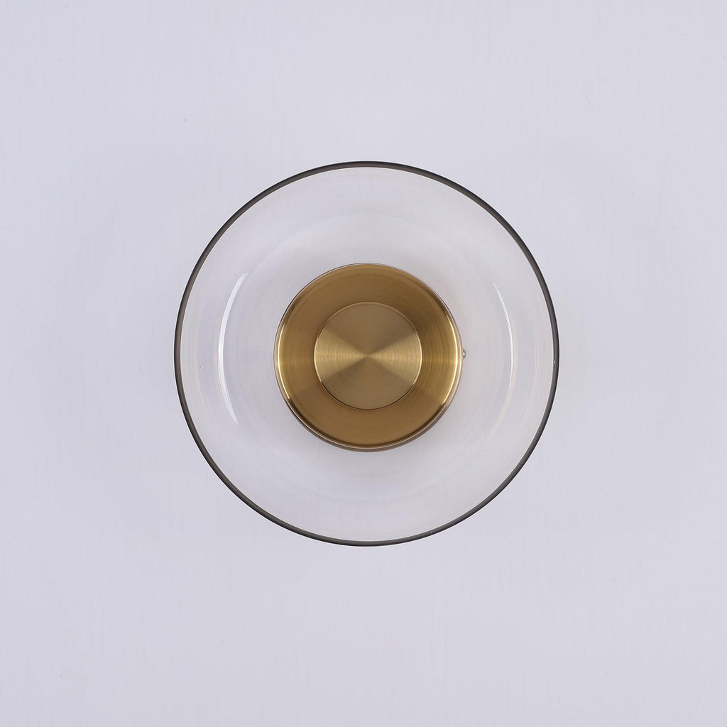 Front view of this Clérachic wall light switched off, with a thin dark glass rim, clear glass and a brushed brass disc.