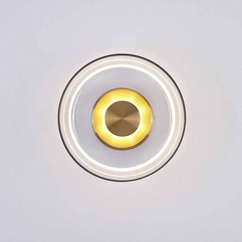 Lit front view of this Clérachic wall light, with a bright ring of light under the clear glass and a glowing brass disc.