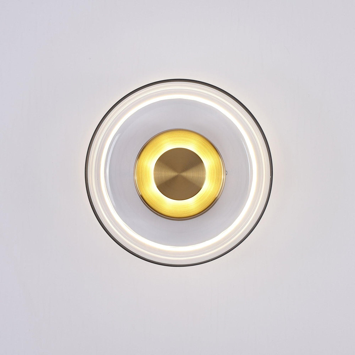 Lit front view of this Clérachic wall light, with a bright ring of light under the clear glass and a glowing brass disc.