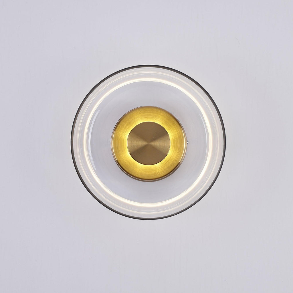 This Clérachic wall light with a clear glass dome, lit, shows a soft white ring and a glowing gold centre on a pale grey wall.