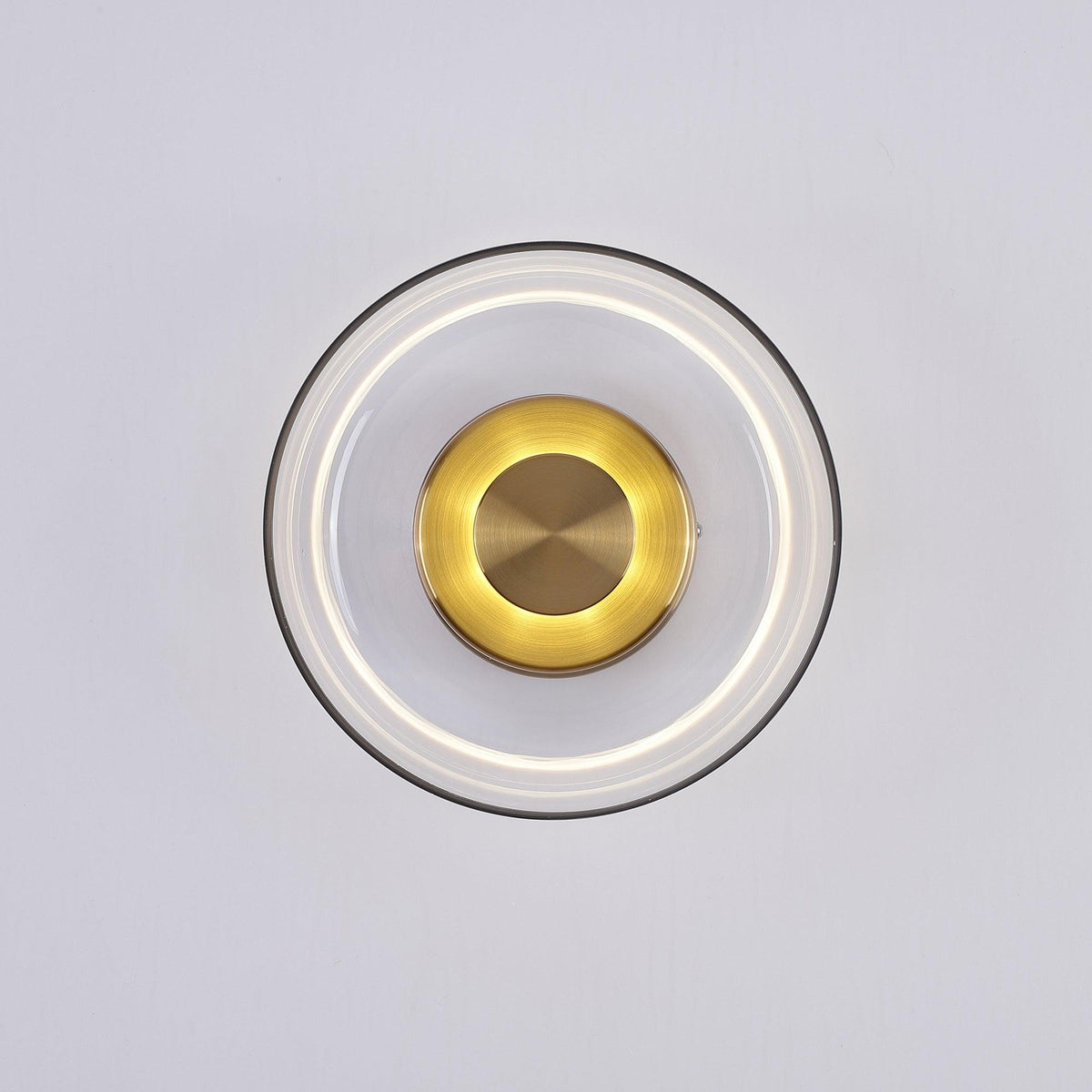 This Clérachic wall light with a clear glass dome, lit, shows a soft white ring and a glowing gold centre on a pale grey wall.