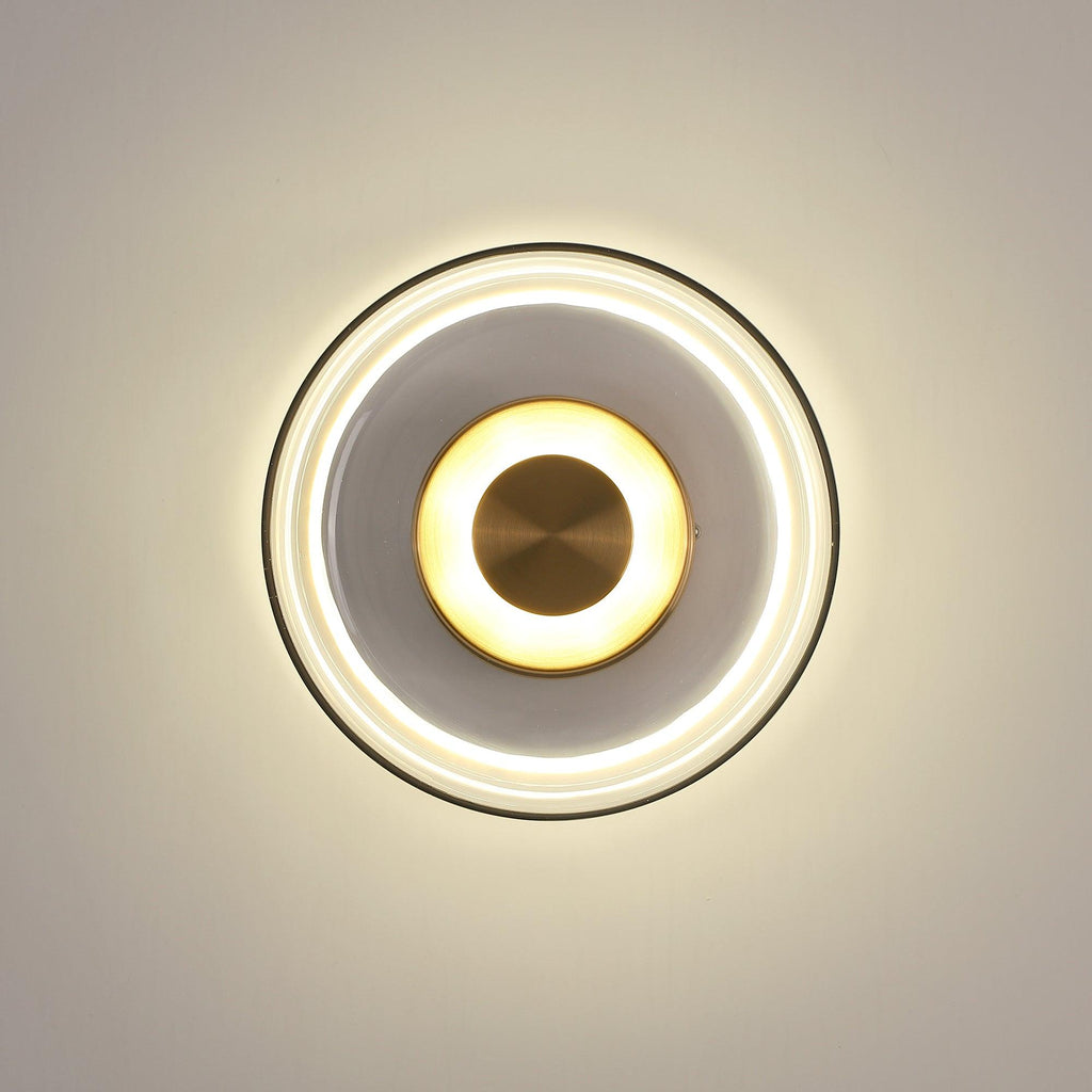 A Clérachic wall light on a cream wall, with a wide halo of warm light and a smoky glass dome around a glowing brass disc.