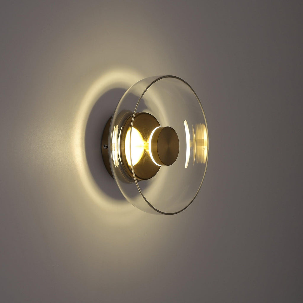 Side view in a dim room: this Clérachic wall light lights its clear glass dome and brass plate from inside and throws a halo behind.