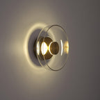 Side view in a dim room: this Clérachic wall light lights its clear glass dome and brass plate from inside and throws a halo behind.