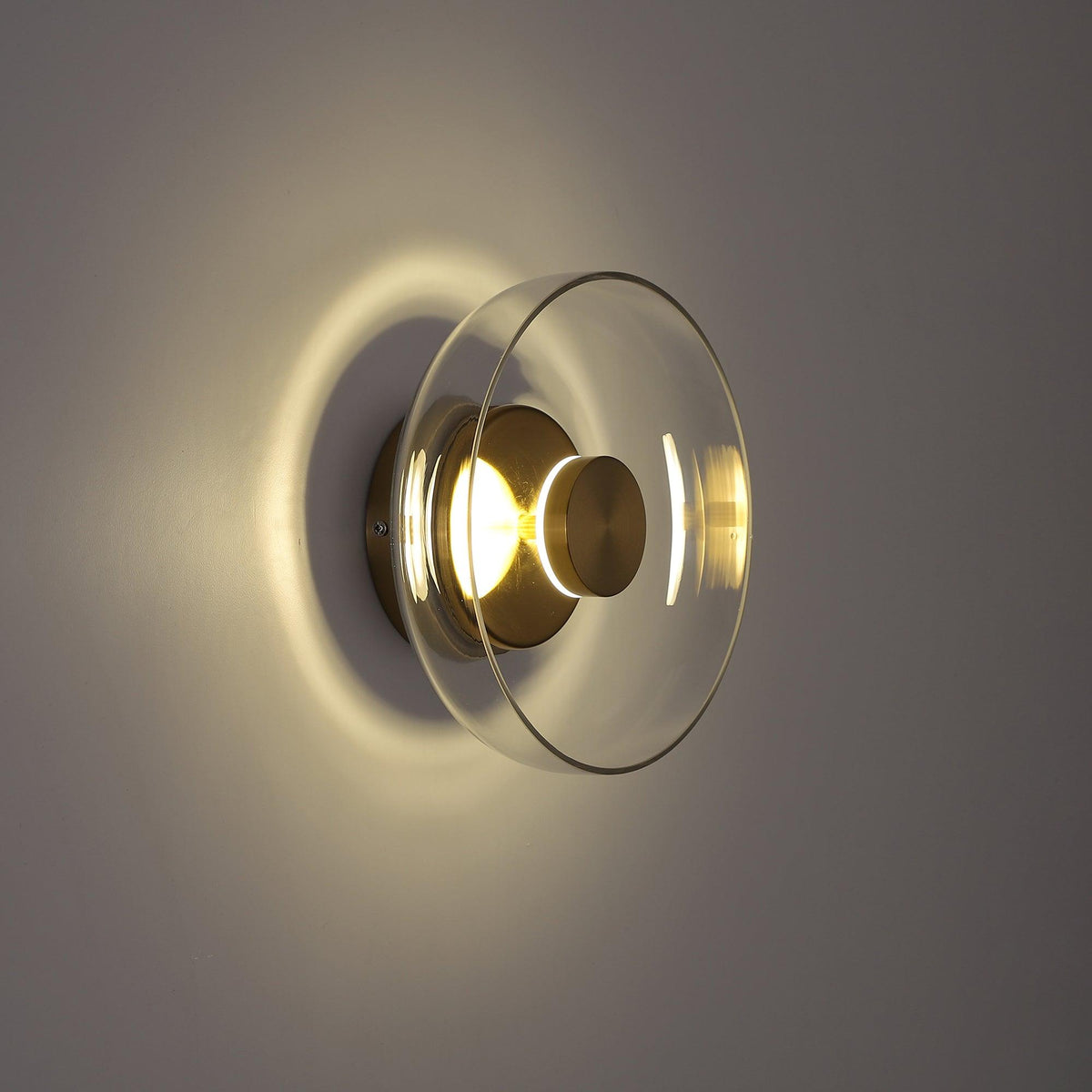 Side view in a dim room: this Clérachic wall light lights its clear glass dome and brass plate from inside and throws a halo behind.