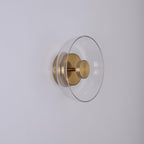 Side view of this Clérachic wall light switched off, with a clear glass dome, a brass stem and a round brass wall plate.