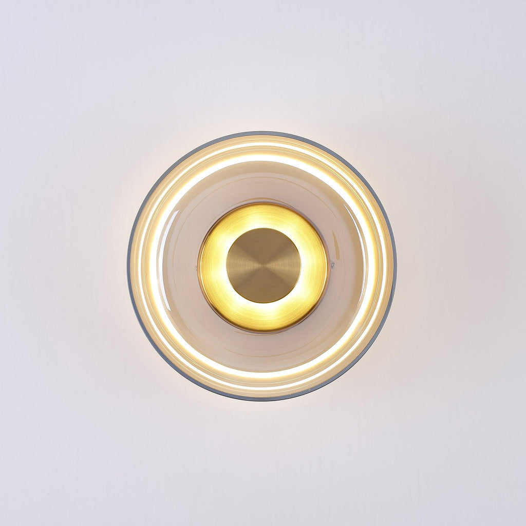 Lit front view of an amber glass Clérachic wall light, with a bright glowing ring beneath the glass and a brass centre disc.