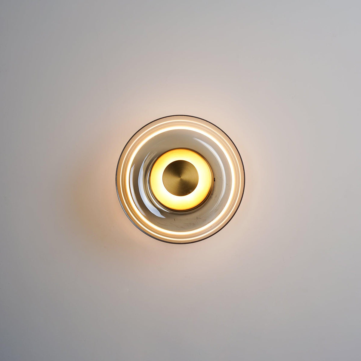 A Clérachic wall light glowing on a grey wall, with a warm centre ring and a clear glass dome edged in a thin dark rim.