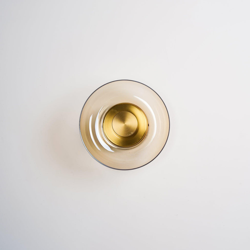 This Clérachic wall light has a pale amber glass dome and a polished brass centre disc, shown unlit on a light wall.