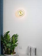 A small Clérachic wall light glows on a white wall above a leafy plant and a row of books beside a blue curtain.