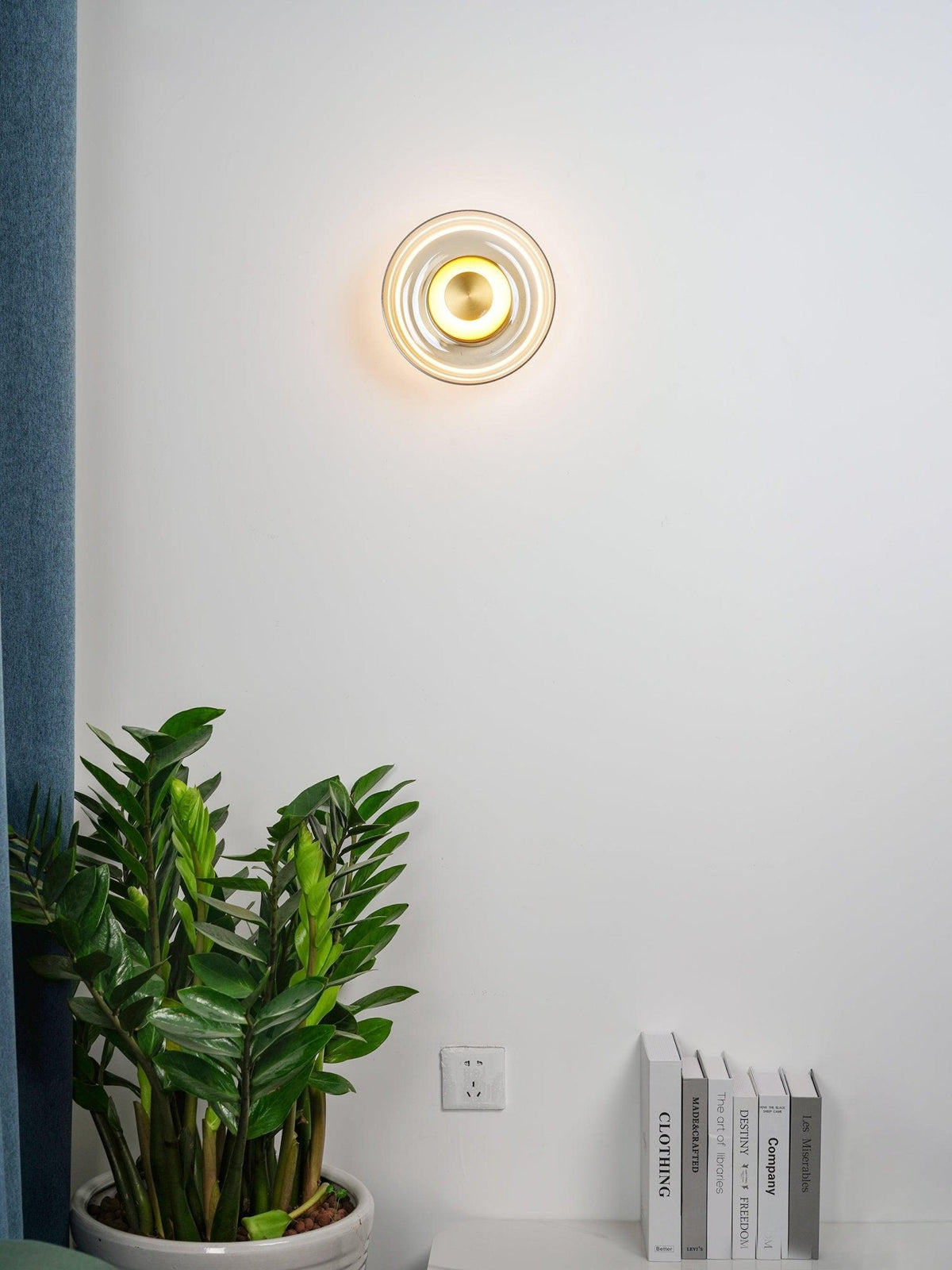 A small Clérachic wall light glows on a white wall above a leafy plant and a row of books beside a blue curtain.