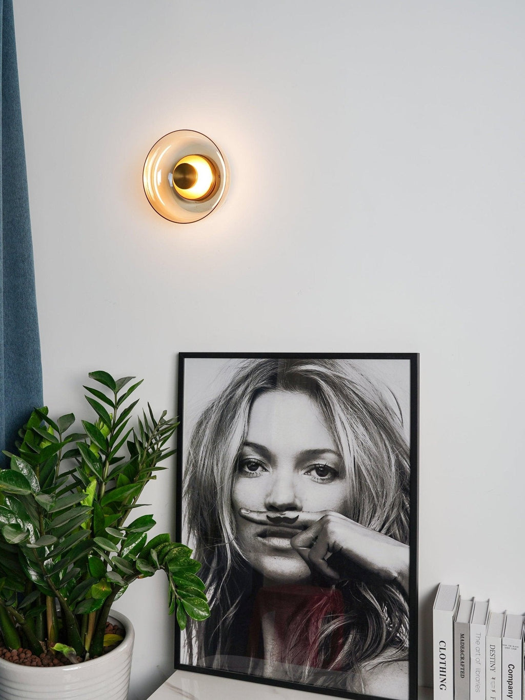 A Clérachic wall light with a warm glow hangs on a white wall above a large potted plant and a framed black-and-white portrait.