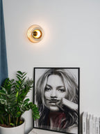 A Clérachic wall light with a warm glow hangs on a white wall above a large potted plant and a framed black-and-white portrait.