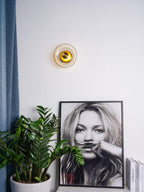 This Clérachic wall light hangs on a white wall above a framed black-and-white portrait, a green plant and a blue curtain.