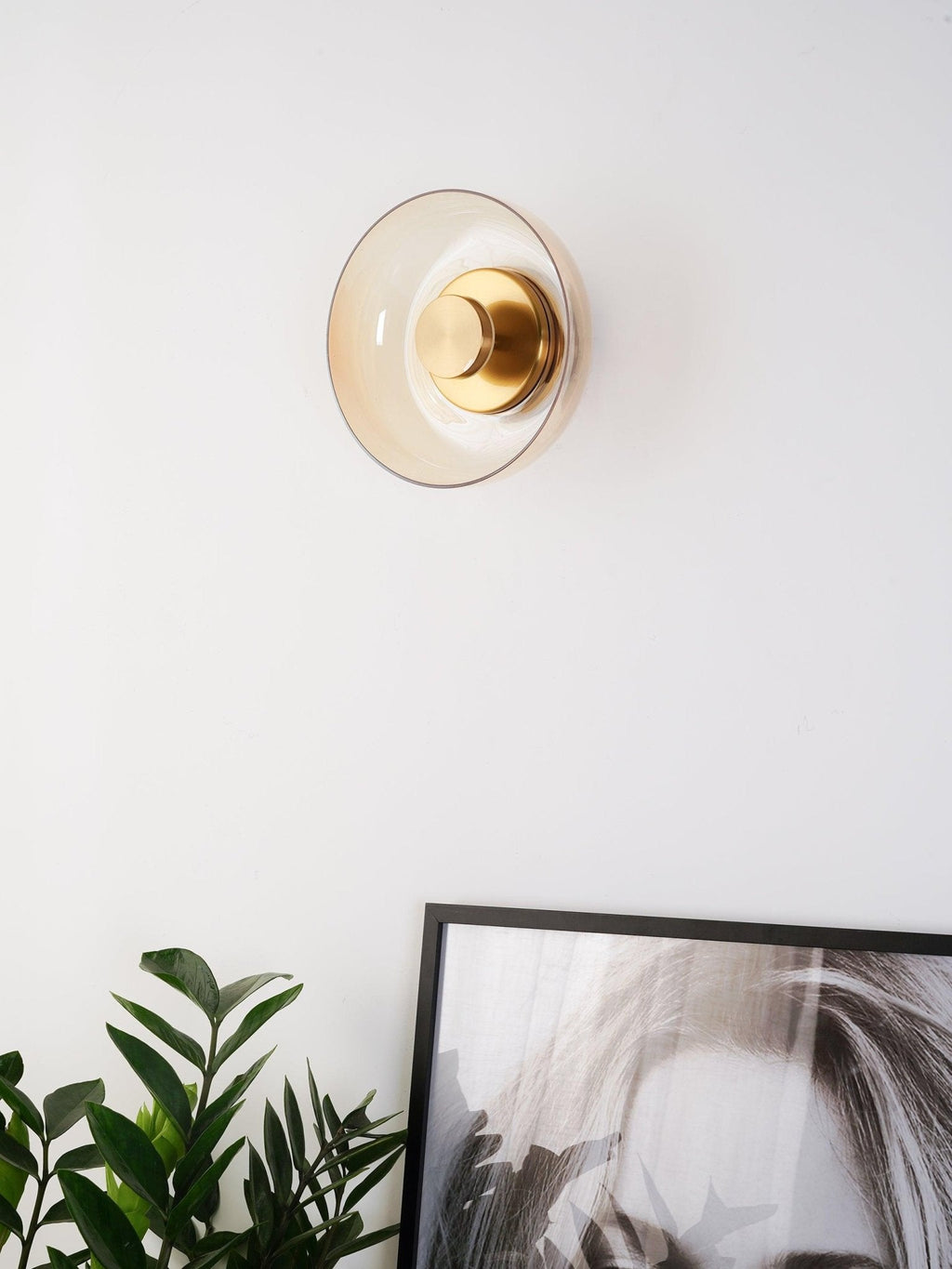 An amber glass Clérachic wall light on a white wall, its brass centre knob catching the light above a plant and a framed print.