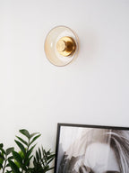 An amber glass Clérachic wall light on a white wall, its brass centre knob catching the light above a plant and a framed print.