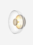 Angled view of this Clérachic wall light on white, with a clear glass dome and a brass disc over a softly glowing interior.