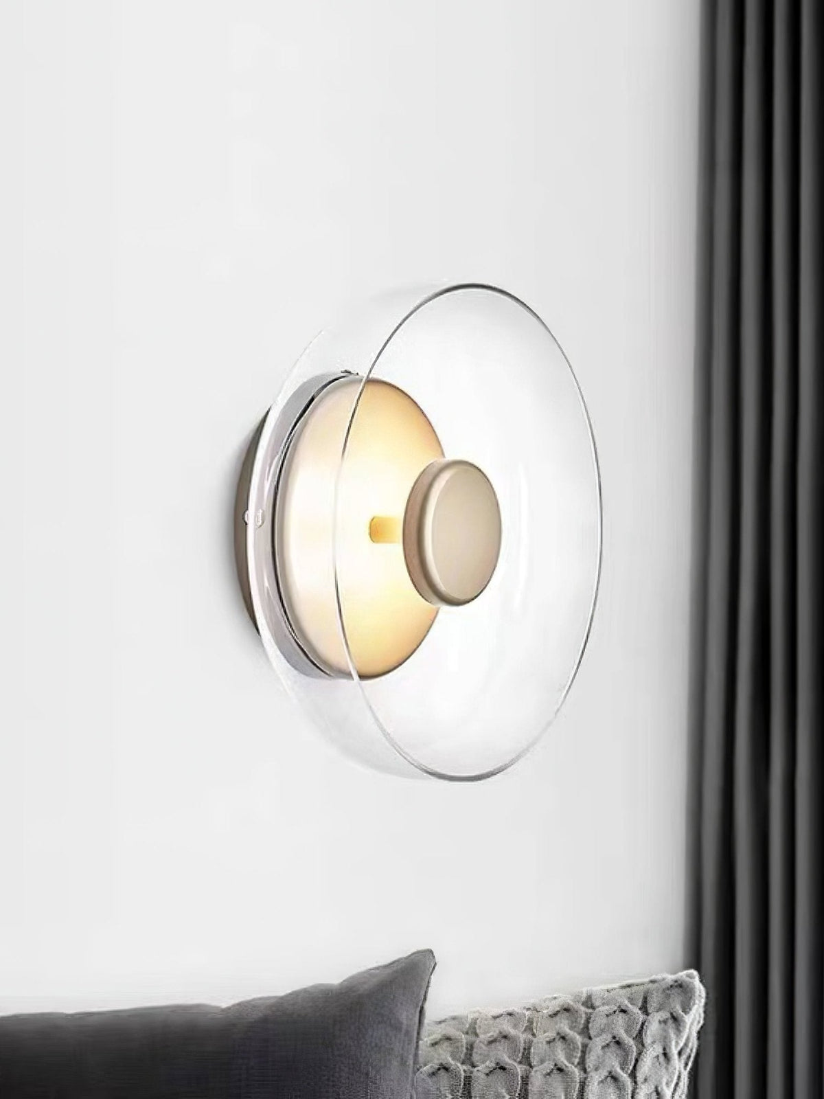 A Clérachic wall light on a white wall beside a dark grey curtain, with a clear glass dome and a small brass centre knob.