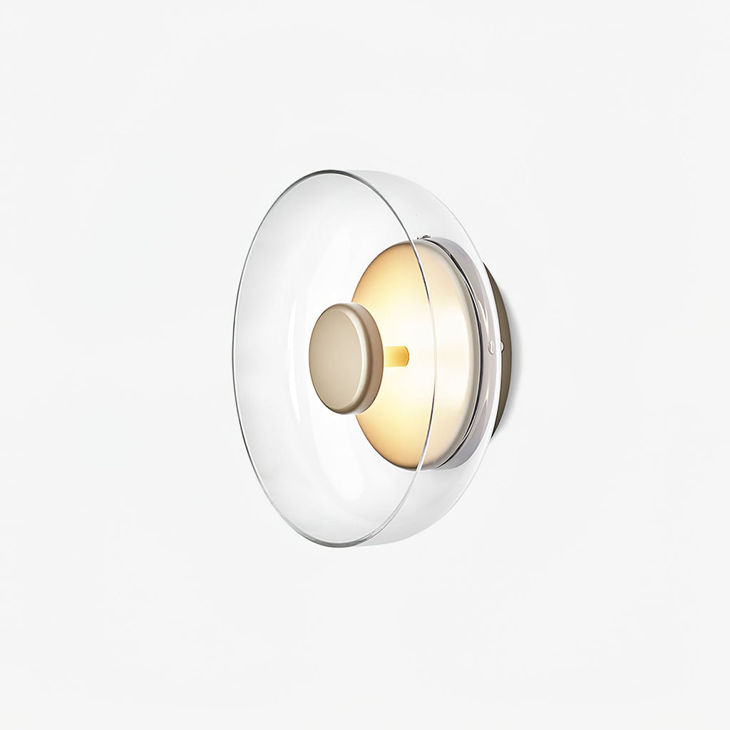 A Clérachic wall light on a very pale background, seen from the side with a clear glass dome and a softly lit brass disc.