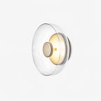A Clérachic wall light on a very pale background, seen from the side with a clear glass dome and a softly lit brass disc.