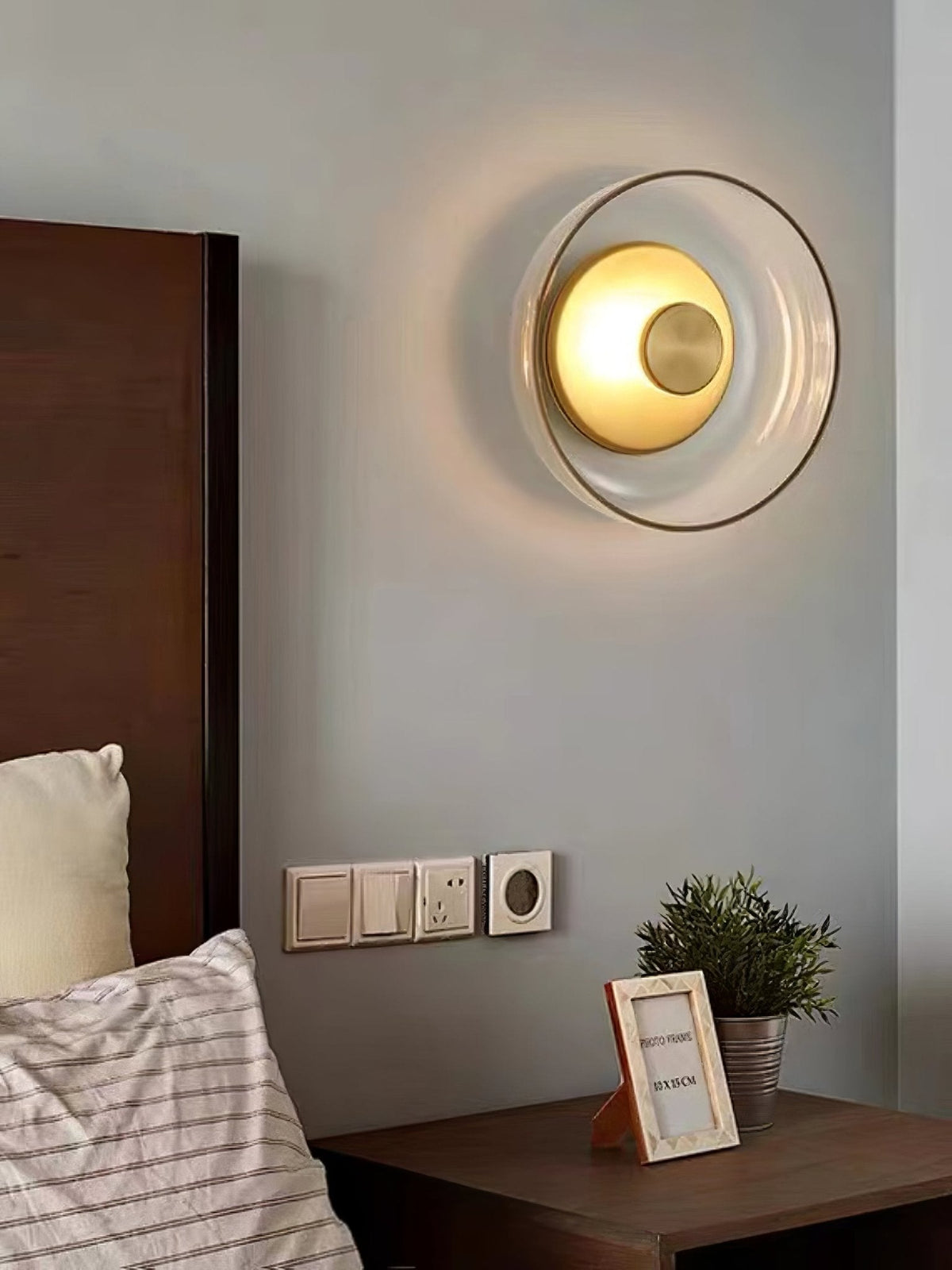 Over a bed with a walnut headboard, this Clérachic wall light glows warm inside its clear glass dome, above a small framed photo.
