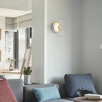 A Clérachic wall light on a pale wall in a bright living room, above a grey sofa with blue and red cushions.