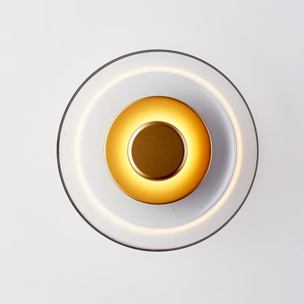 Close front view of this Clérachic wall light lit, with a bright inner ring, a gold-toned centre disc and a clear glass dome.