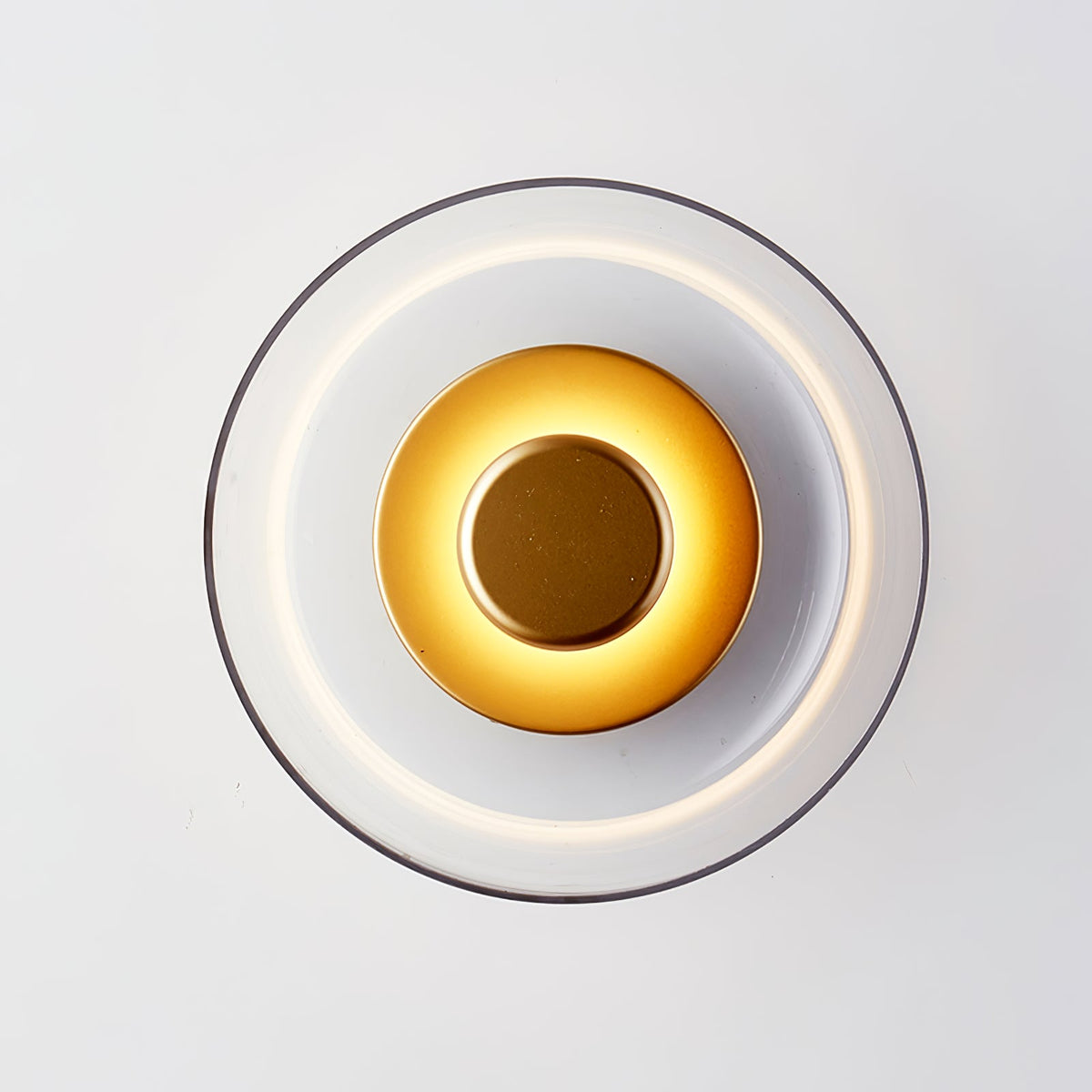Close front view of this Clérachic wall light lit, with a bright inner ring, a gold-toned centre disc and a clear glass dome.