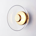 Close view of this Clérachic wall light with a clear glass dome and a polished brass disc, tilted slightly on a pale wall.