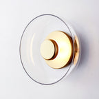 Close view of this Clérachic wall light with a clear glass dome and a polished brass disc, tilted slightly on a pale wall.