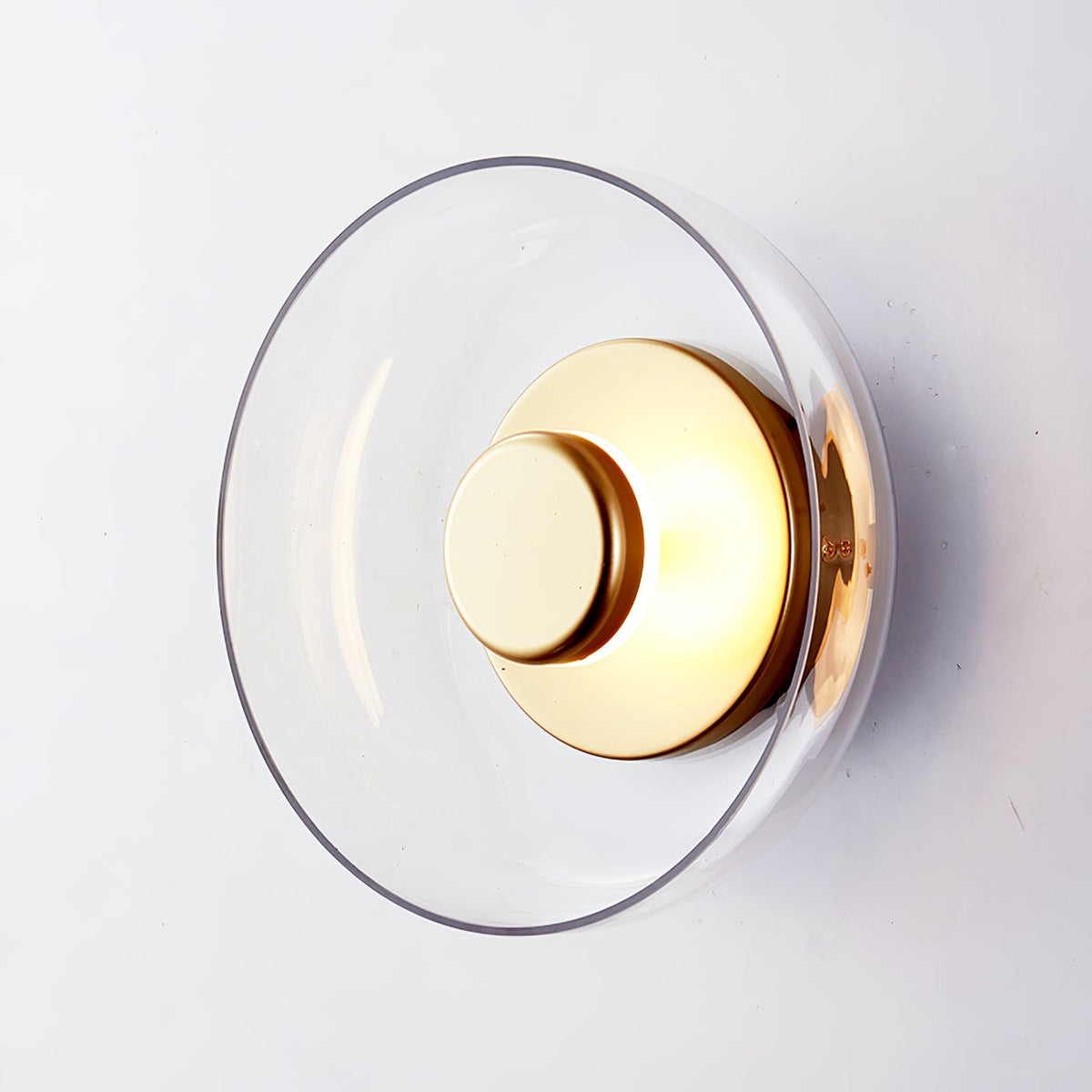 Close view of this Clérachic wall light with a clear glass dome and a polished brass disc, tilted slightly on a pale wall.