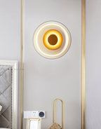 A Clérachic wall light glows on a pale wall between two brass-framed panels, above a wall socket and a small gold ornament.