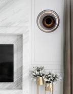 A smoke grey glass Clérachic wall light with a black body glows beside a grey curtain, above a marble fireplace and gold vases.