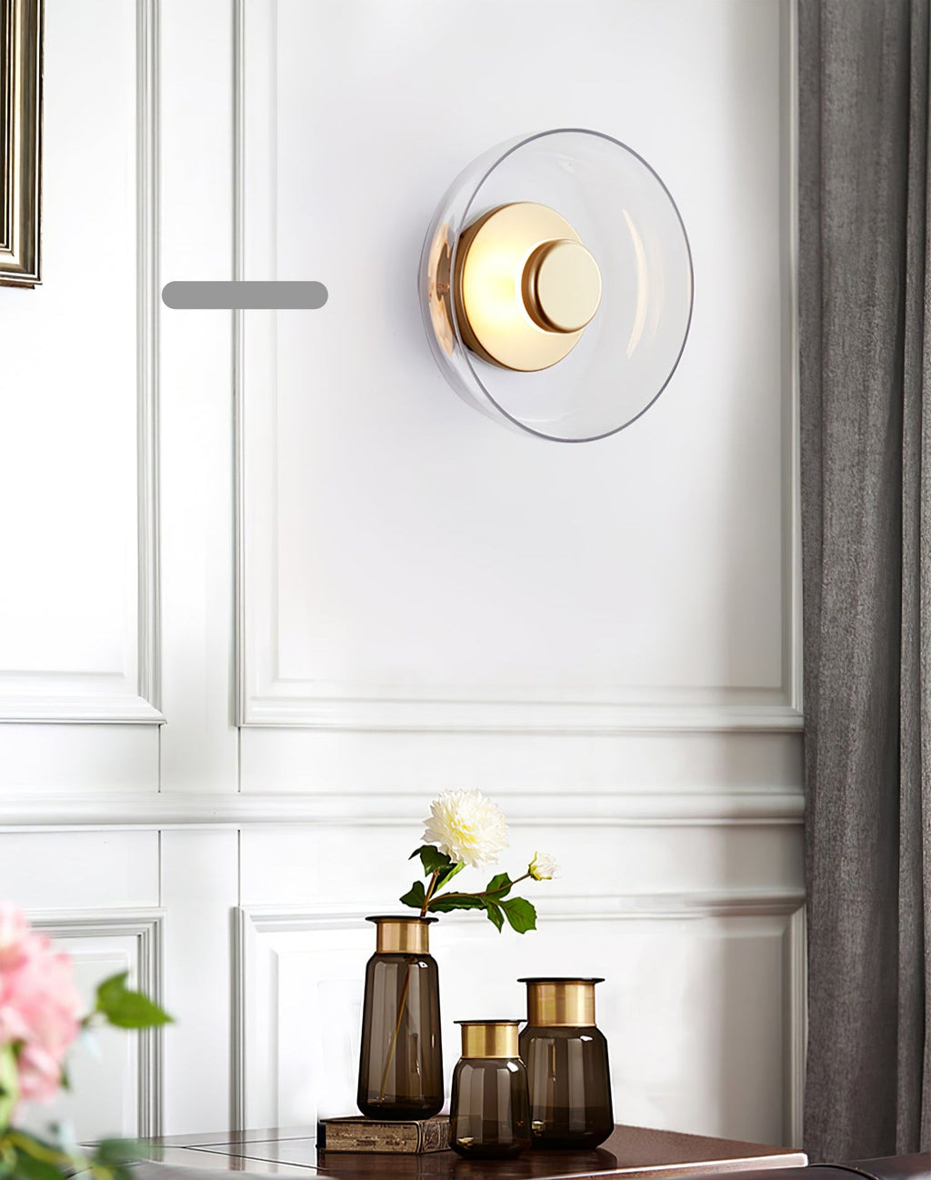 A Clérachic wall light with a clear glass dome and a brass disc hangs on a panelled wall above a console with brown glass vases.