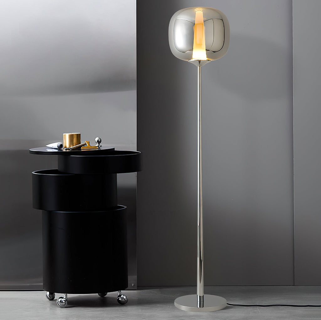 Next to a black drum side table, a chrome Clérachic floor lamp with a smoked glass head stands against a grey wall.