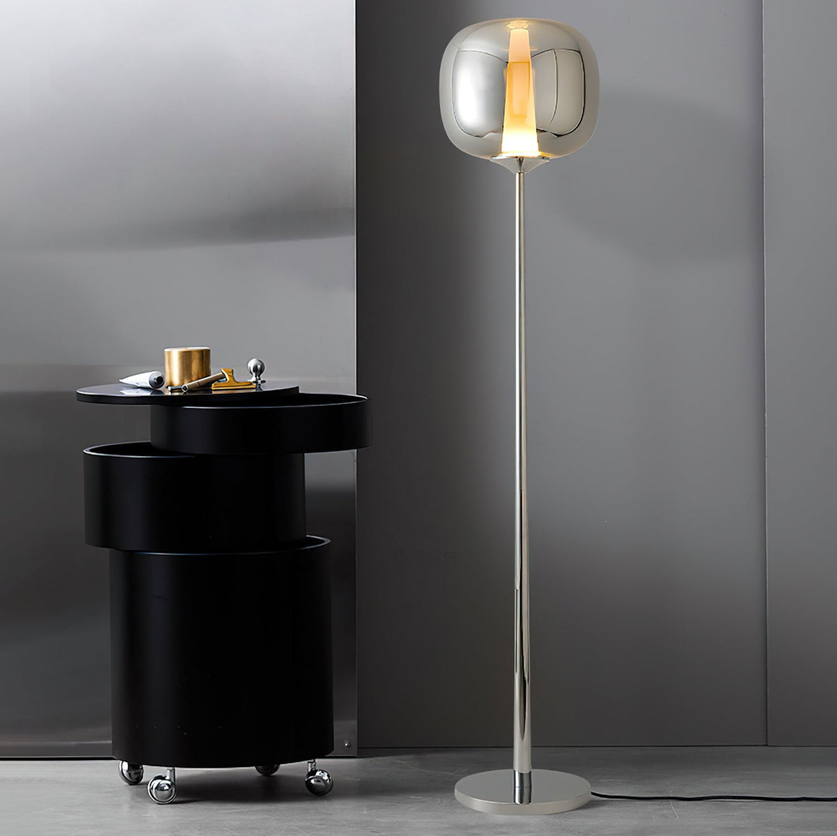 Next to a black drum side table, a chrome Clérachic floor lamp with a smoked glass head stands against a grey wall.