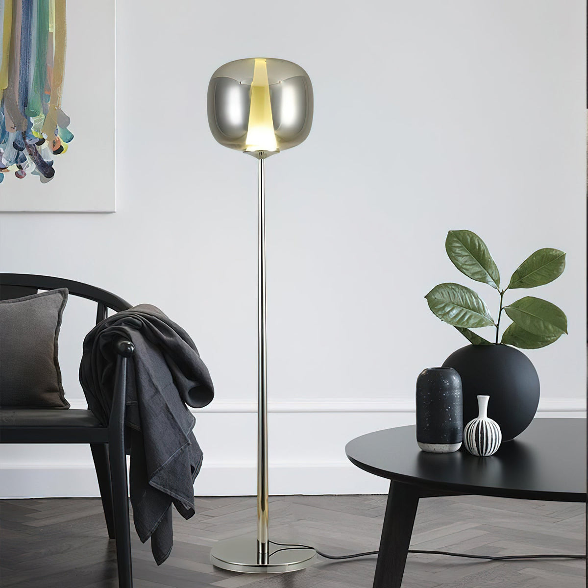 Chrome Clérachic floor lamp with a smoked glass head beside a black round table, a dark chair and a vase of leaves.