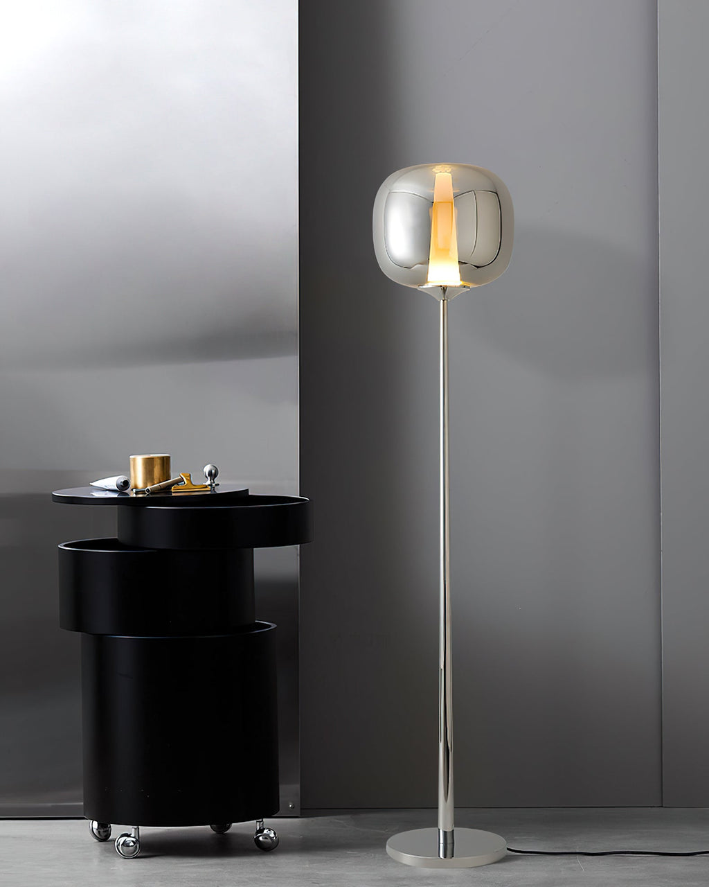 A chrome Clérachic floor lamp stands tall beside a black drum table with brass objects on top, against a grey wall.