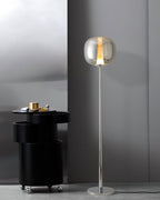 A chrome Clérachic floor lamp stands tall beside a black drum table with brass objects on top, against a grey wall.