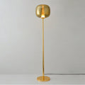 Gold Clérachic floor lamp lit warm, its gilt glass head glowing, standing on a round gold base against grey.